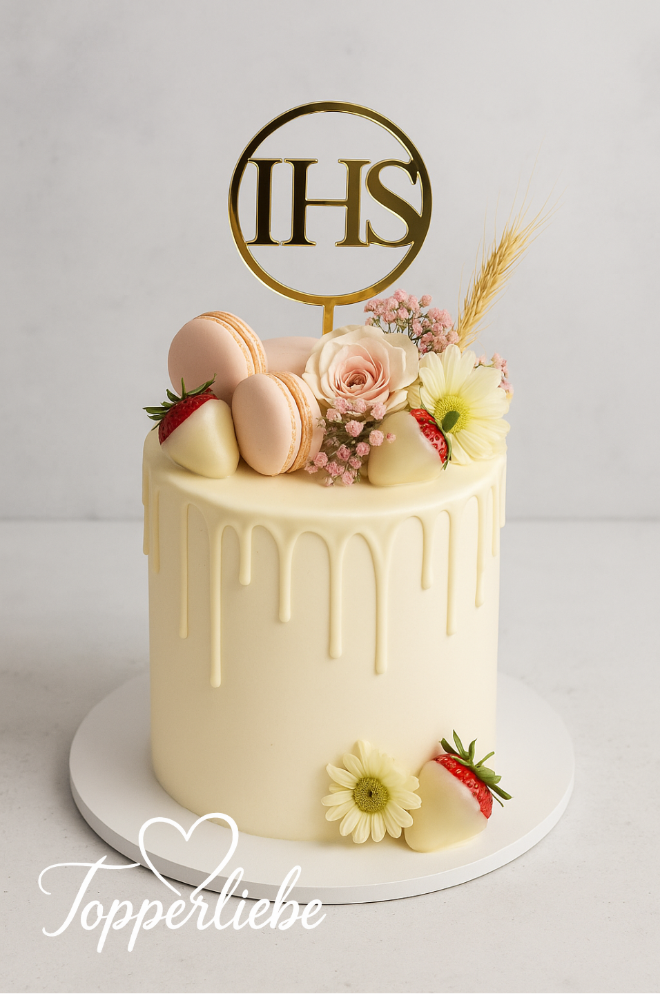 Weißer Kuchen mit goldenem IHS-Topper aus Acryl, dekoriert mit Blumen, Makronen und Erdbeeren.
