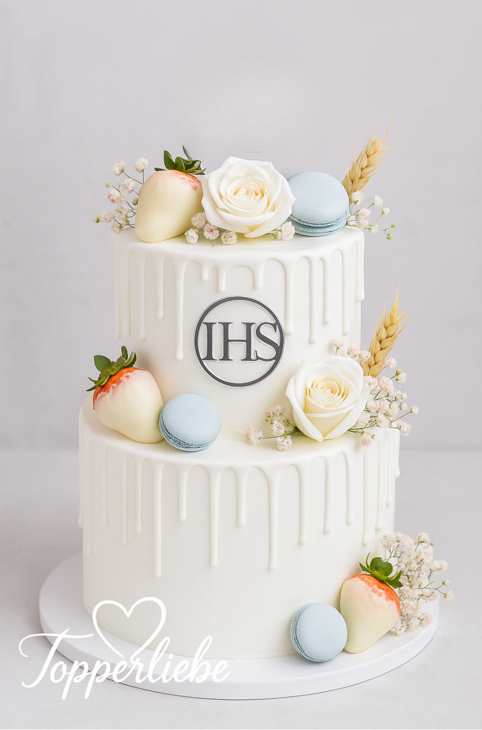 Weißer zweistöckiger Kuchen mit seitlichem IHS-Dekor aus Acryl, dekoriert mit weißen Blumen, Makronen und Ähren